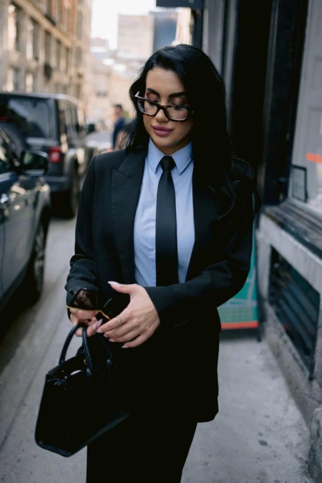Stéphanie Nachar en tailleur noir et cravate, marchant dans une rue de Montréal avec son sac à main.
